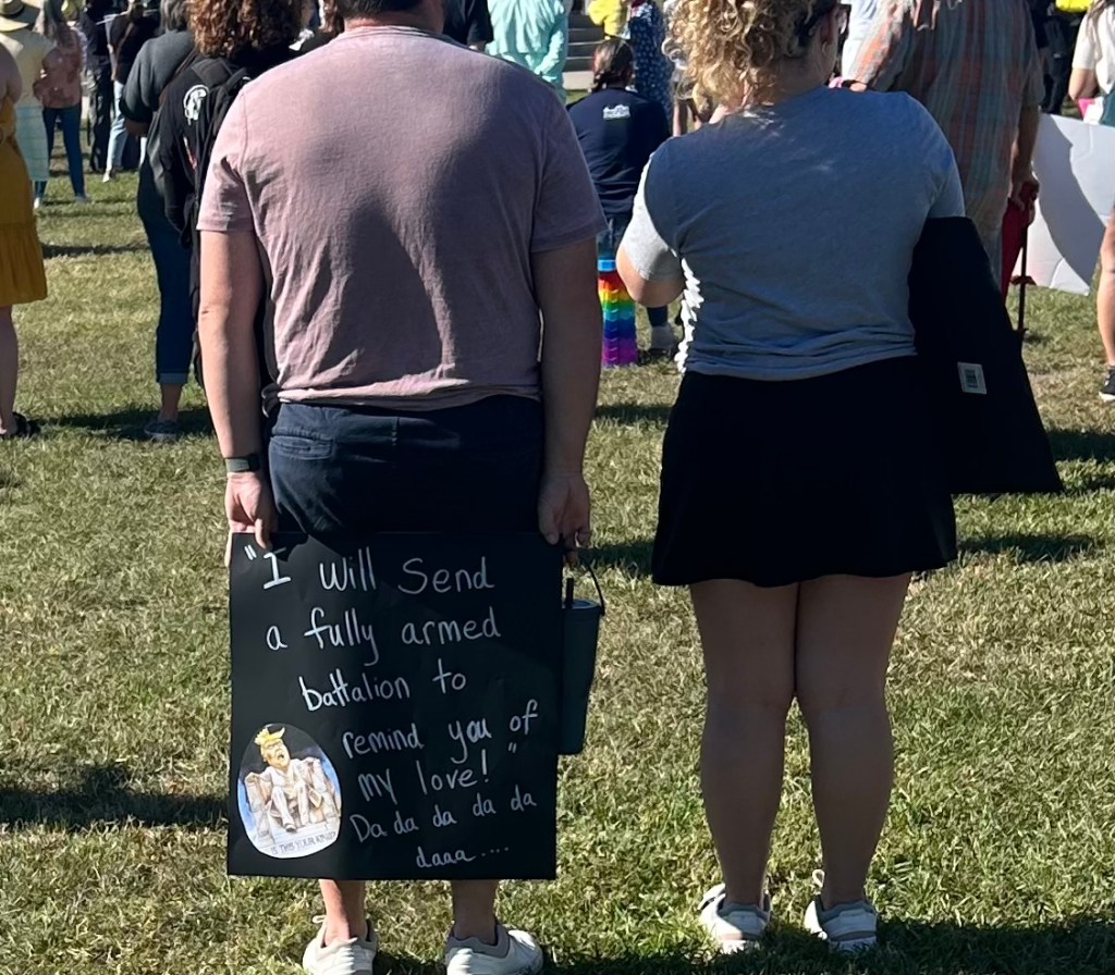 Protestor with sign reading "I will send a fully armed battalion to remind you of my love! Da d da da da daaa"